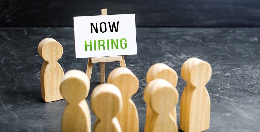 Little people-shaped wooden block are gathered around a wood easel with a sign reading Now Hiring.